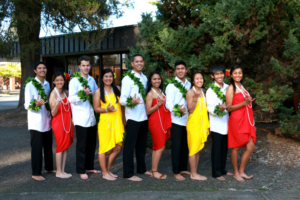 Alternating men and women in the Hawaiian club.  Women in alternating red and yellow dresses, men in white shirts, black pants, and have sashes of garlands.  