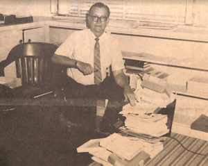A man, Milo Ross, is on one knee in an office organizing books and papers. 