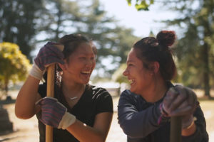 Two girls look at each other, laughing, while wearing gloves and using shovels in an outdoor clearing. 