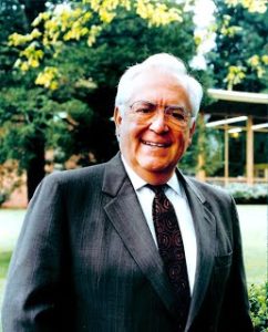 Color photograph, David LeShana in a suit on the George Fox College campus. Grass, trees, and a building in the background. 