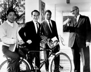 Four men, one of whom is on a bike, pose for a photograph on the George Fox College Campus. Black and white photograph. 